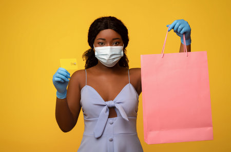 Young black woman in face mask and rubber gloves holding shopper bag with mockup and credit card on...