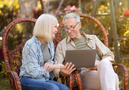 Happy elderly couple relaxing in wicker chairs in open air and using laptop computer, copy space