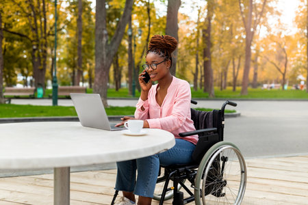 Disabled black lady in wheelchair using laptop, having conversation on smartphone at outdoor cafe in...