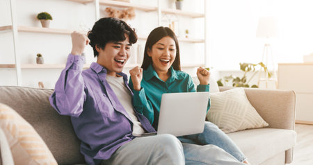 Emotional Asian couple with laptop making YES gesture, celebrating success, sitting on couch at home, free spaceの写真素材