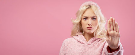 Young Woman in Pink Sweater Gestures to Show Stop Sign During Photo Shoot on Pink Backgroundの写真素材