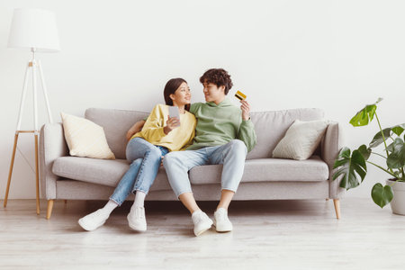 Cheerful young Asian couple sitting on sofa with credit card and cellphone, shopping online in living room, copy spaceの写真素材