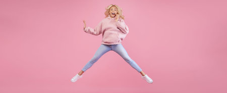 Girl Jumps in Excitement Against a Pink Background While Wearing a Pink Hoodie and Jeansの写真素材