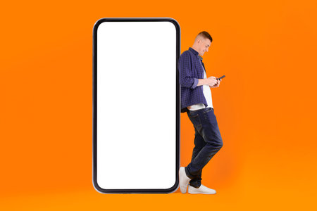 Millennial Man Using Phone Leaning On Huge Smartphone, Yellow Background