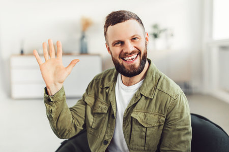 Joyful Man Waving Hand Smiling To Camera Sitting At Homeの写真素材