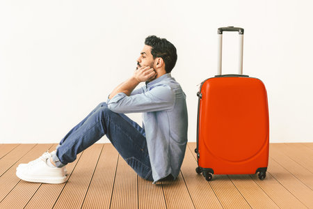 Upset male tourist sitting near suitcase and looking away at copy space, waiting for flight over light wallの写真素材