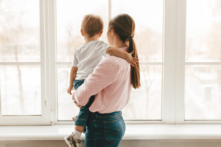 Young mother holding her little son on hands, standing near window and looking through it, back...