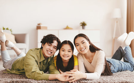 Cheerful millennial asian lady, guy and teen daughter lie on floor and hold hands in living room interiorの写真素材