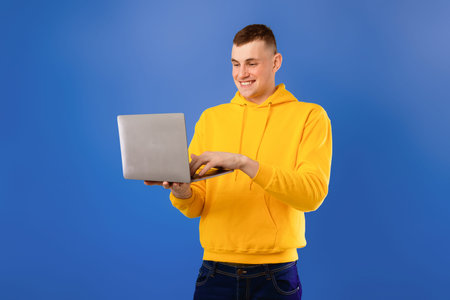 Happy caucasian man enjoying his new modern laptop, typing on keyboard and smiling, blue background,...