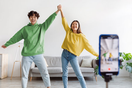 Happy young Asian couple making video on cellphone, dancing in front of camera at homeの写真素材
