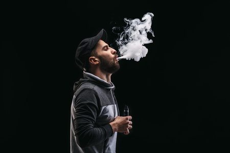 Man Blows Vapor From Vape While Standing in Dark Space With Smoke Rising Upward