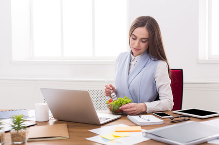 Office Worker Eating Salad While Using Laptop During Lunch Break in Bright Workspaceの写真素材