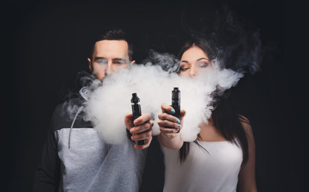 Two People Hold Vape Devices and Exhale Smoke in a Dark Setting