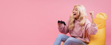 Young Woman Plays Video Game on a Yellow Bean Bag Against a Pink Background With Joy and Excitementの写真素材
