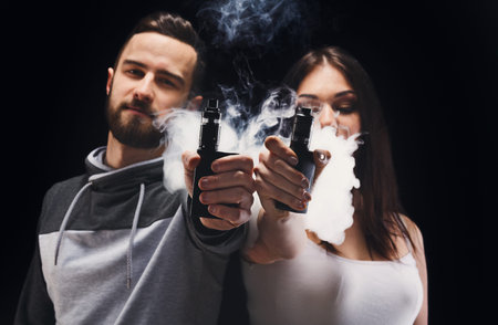 Couple Holding Vape Pens and Exhaling Vapor in a Dark Setting During an Indoor Activity