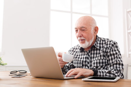 Elderly Man Drinks Coffee While Working on Laptop in Bright Room With Natural Light