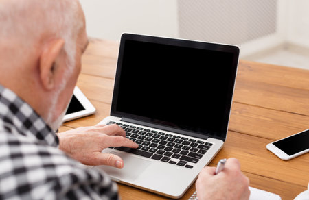 Senior Man Working on Laptop and Taking Notes at a Wooden Table During Daytime