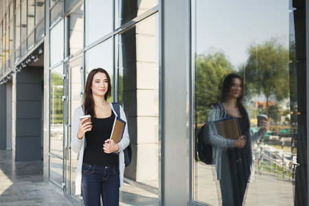 A Young Woman Stands Outside a Modern Building, Holding a Coffee Cup and a Notebook