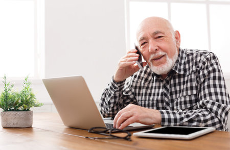 Senior Man Talks on Phone While Working at Laptop in Bright Room With Tablet Nearby
