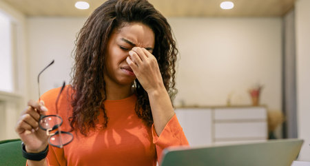 Woman Holds Glasses and Rubs Her Forehead While Looking at a Laptop in a Work Settingの写真素材