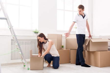 Couple Packs Boxes in New Home During Moving Day in Bright Living Room With Ladder and Plants