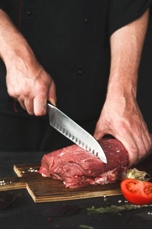 Chef Cuts Fresh Meat on a Wooden Board With a Knife on a Dark Background in a Kitchen Setting