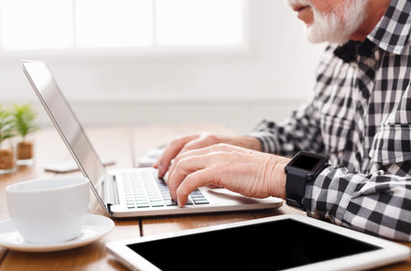 Senior Man Typing on Laptop While Sitting at a Wooden Table With a Cup and Tablet Nearby