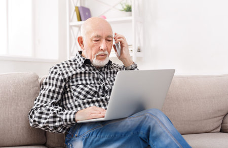 Older Man Talks on Phone While Using Laptop at Home on a Cozy Afternoon