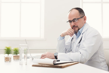 Doctor Working on Laptop at a Desk in a Bright Office Setting With Plants and Notebook
