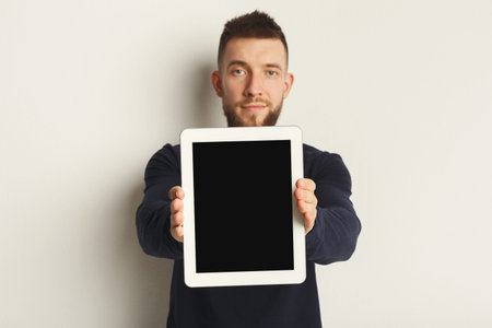 Man Holds Tablet in Front of Him While Standing Against a Plain Background