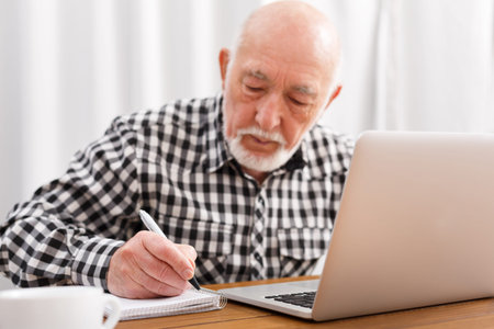 Senior Man Writes Notes While Working on Laptop at Home During Day