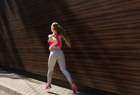 Woman Runs on City Street Near Wooden Wall While Wearing Sports Gear in Daylight