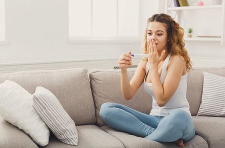 Woman Sits on Couch and Looks Surprised at Pregnancy Test Result in Modern Living Room