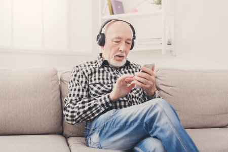 Elderly Man Wearing Headphones Checks Smartphone While Sitting on a Sofa in a Cozy Room