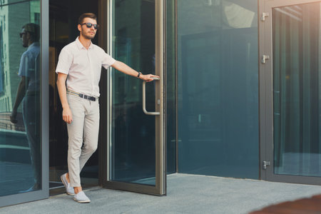 Man Steps out of Modern Building in Urban Area on a Sunny Day Wearing Sunglasses and Casual Attire