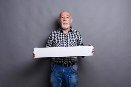 Elderly Man Holding a Blank Sign Against a Gray Background While Wearing Casual Clothing