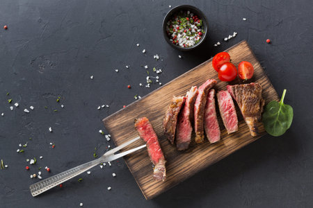 Freshly Cooked Steak Slices Served on a Wooden Board With Tomatoes and Seasoning on a Dark Surfaceの写真素材
