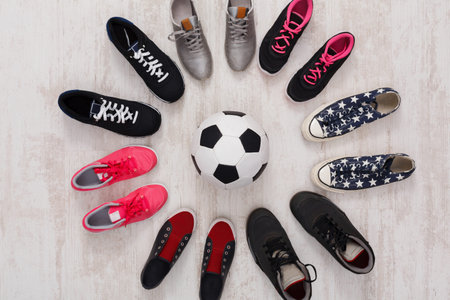 Different Types of Shoes Arranged Around a Soccer Ball on a Wooden Floorの写真素材