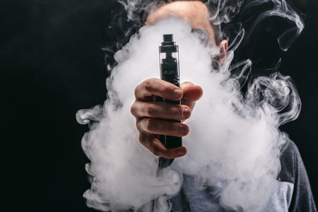 Man Holds Vape Device While Surrounded by Clouds of Vapor in Dark Setting