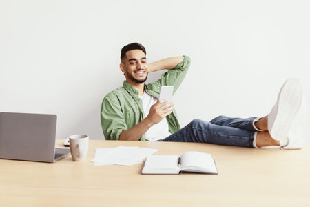 Young Man Relaxing at Desk While Using Smartphone and Enjoying Coffee During Work Hours