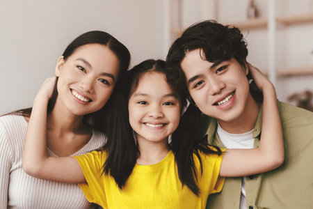 Smiling teenage child hugs young asian mom and dad sits on sofa and looks at camera