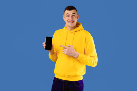 Guy Showing Smartphone Pointing At Blank Screen Over Blue Background