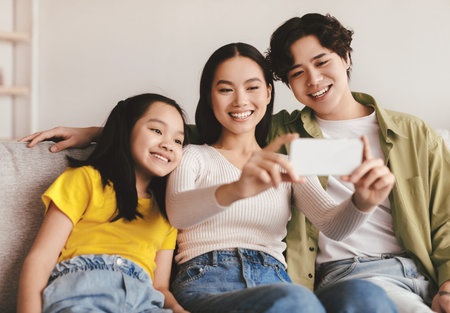 Glad smiling young asian couple and teen daughter making photo on phone on sofa in living room