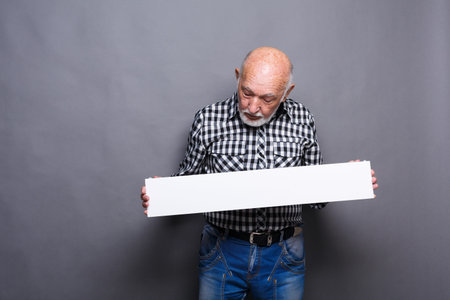 Older Man Holds Blank Sign While Looking Down in Studio With Gray Background