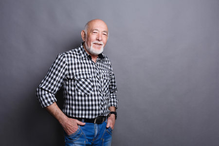 Older Man Poses With a Smile While Wearing a Checkered Shirt and Jeans Against a Gray Background