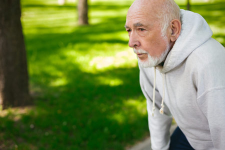 Elderly Man in Gray Hoodie Resting During Exercise in Park on Sunny Dayの写真素材