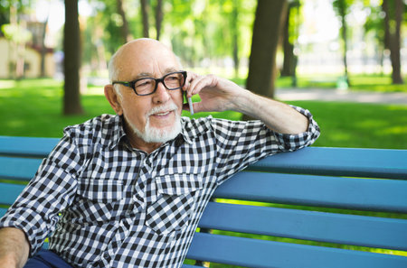 Elderly Man Talks on the Phone While Sitting on a Blue Bench in a Sunny Park