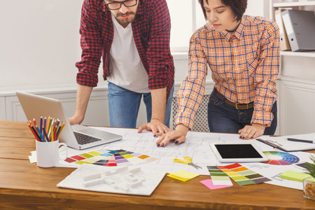 Team Members Collaborate on Design Project at Office Desk With Materials and Laptop