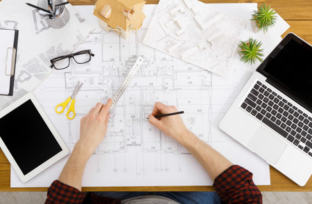 Person Working on Architectural Plans With Tools and Technology on a Wooden Desk