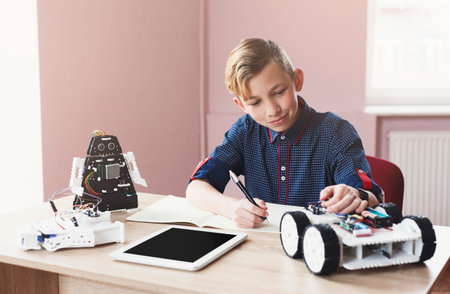 Young Boy Works on Robotics Project at Table With Tablet and Robot Models